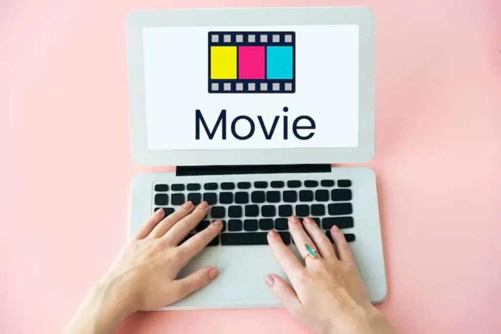 A top-down view of a person's hands typing on a laptop against a pink background, with the screen displaying a colorful clapperboard icon and the word 'Movie'.